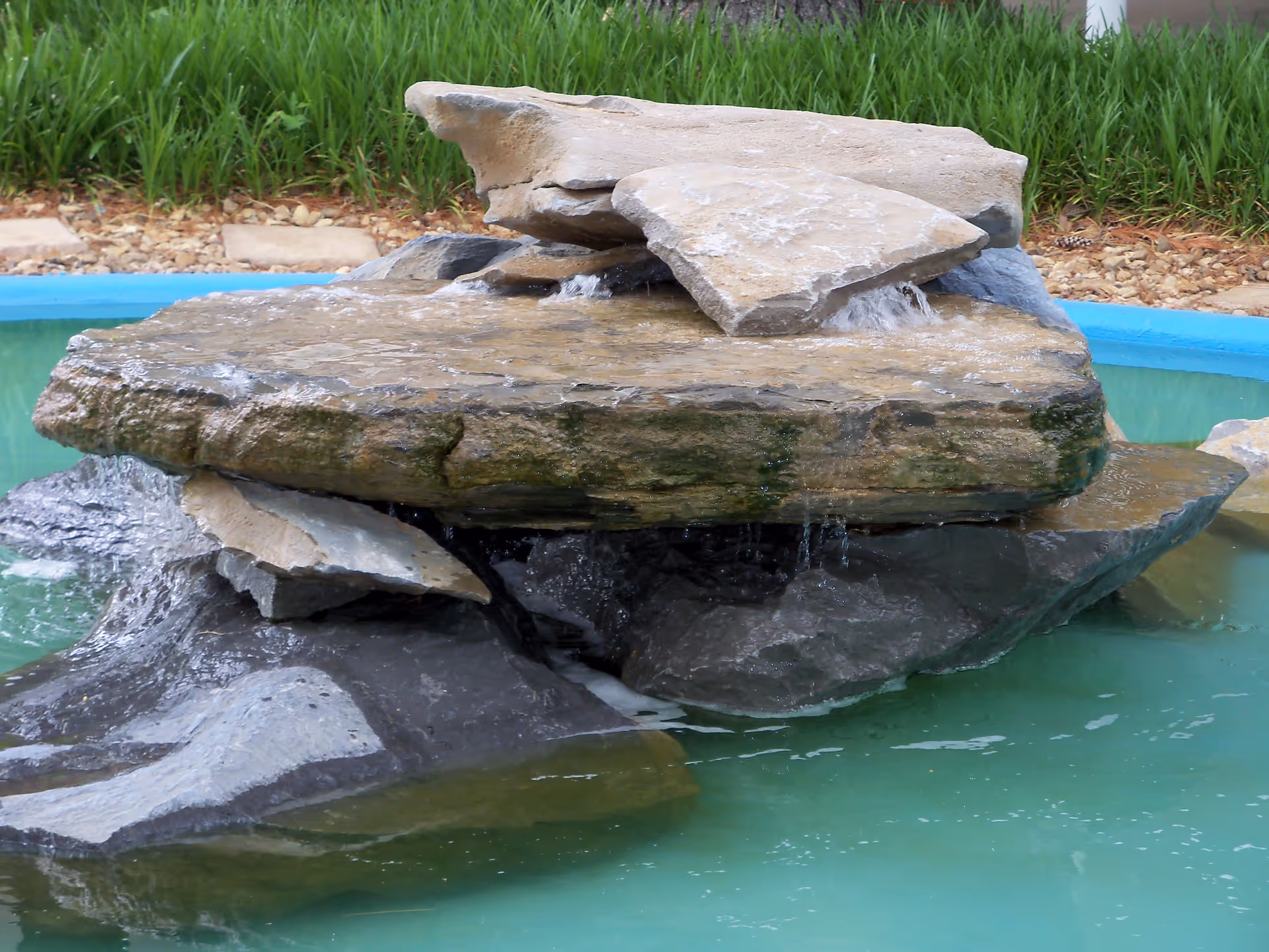 A close-up view of a small rock waterfall feature with water gently flowing over layered flat stones into a turquoise pool, surrounded by green plants and small rocks.