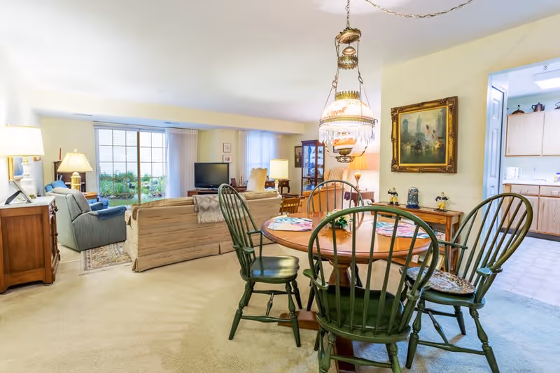 A cozy living and dining area in a senior living facility. The foreground features a round wooden dining table with four green wooden chairs and a decorative hanging lamp above. The background shows a living room with a beige sofa, a blue armchair, a TV on a stand, and large windows with vertical blinds letting in natural light. To the right, there is a view into a kitchen with wooden cabinets and white appliances. The walls are decorated with framed artwork and lamps provide warm lighting.