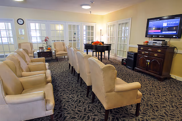 A common area with two rows of beige upholstered chairs facing a television mounted on the wall. There is a wooden cabinet beneath the TV, a black grand piano with a flower arrangement on top, and glass-paneled doors along the walls. The room has patterned carpet and soft yellow walls.