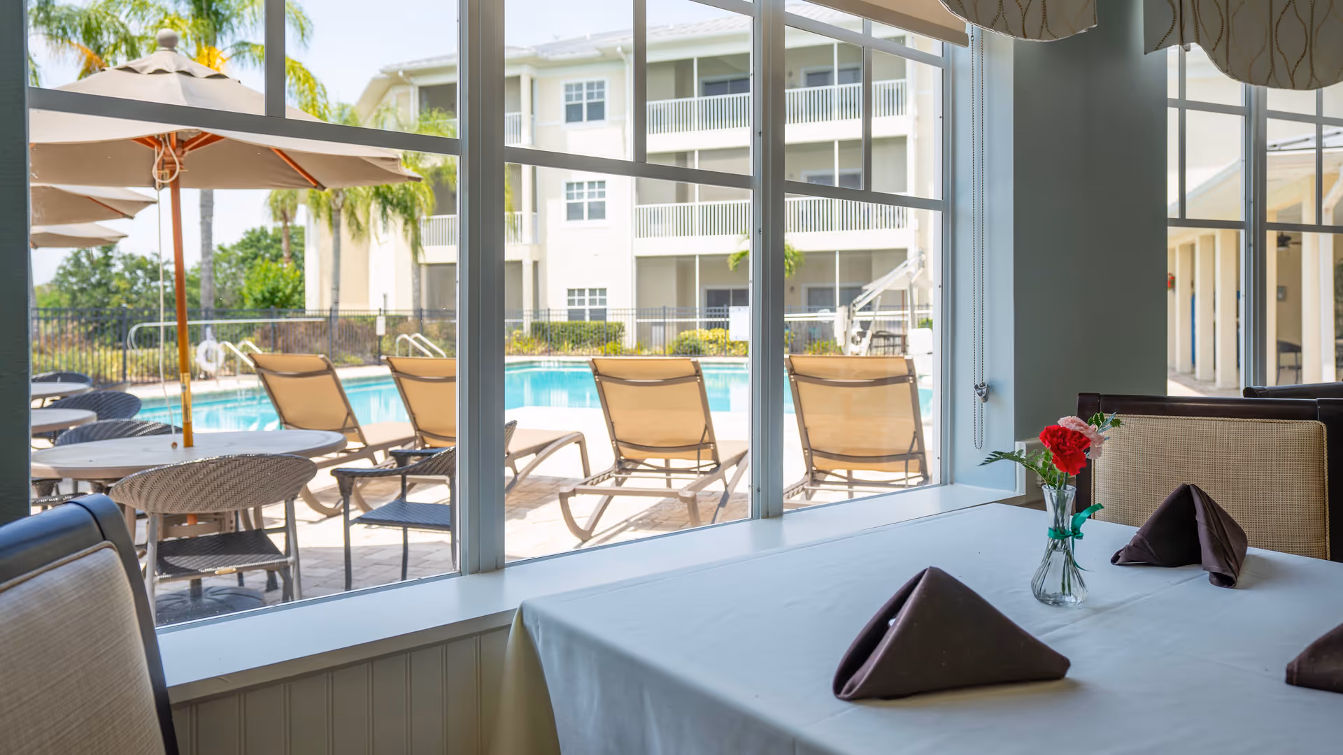 View from inside a dining area looking out through large windows at an outdoor pool area with lounge chairs and umbrellas. A table with a white tablecloth, folded brown napkins, and a small vase with red and pink flowers is visible in the foreground.