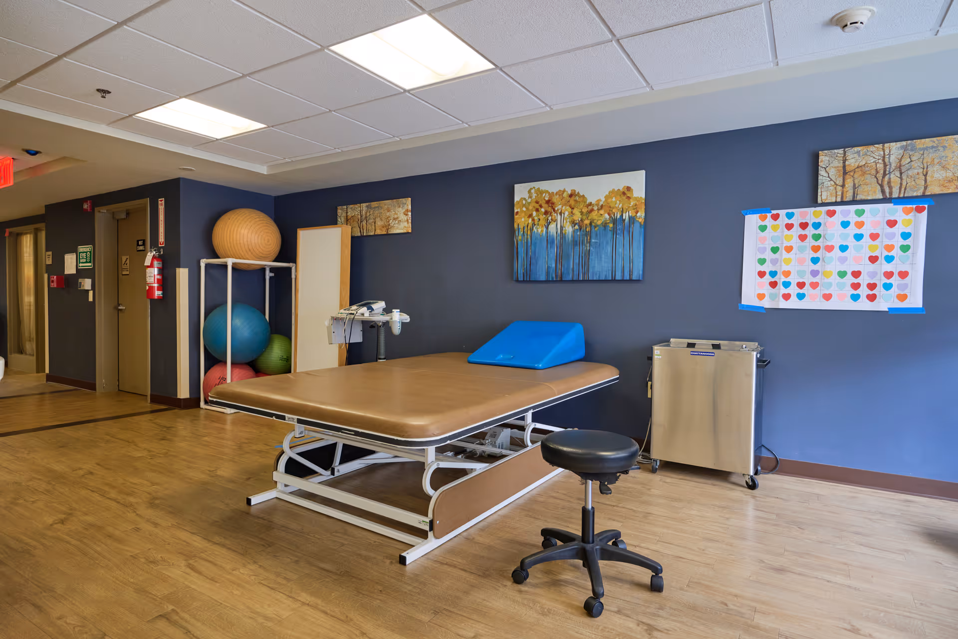 A rehabilitation room with a brown therapy table, a black rolling stool, and exercise balls stored on a white rack against a dark blue wall. The room has wooden flooring, a ceiling with fluorescent lights, and colorful artwork and a chart with heart shapes on the wall.