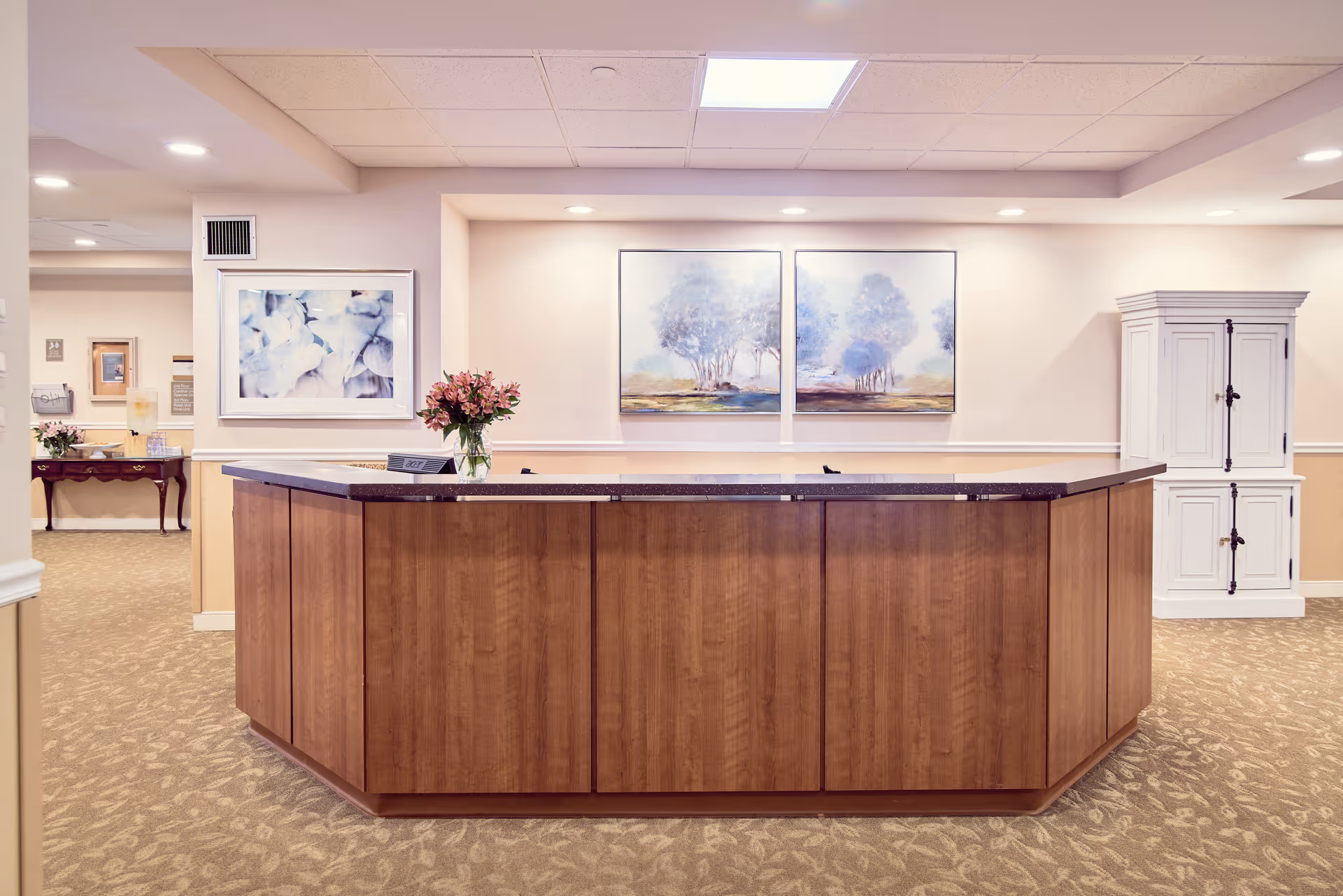 Reception desk area in a senior living facility with a wooden counter, a vase of pink flowers, and three framed paintings on the wall behind it. The space is well-lit with ceiling lights and has beige walls and carpeted flooring. A white cabinet is visible on the right side and a hallway with additional furniture and decor is seen to the left.