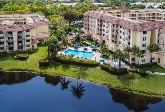 Aerial view of The Horizon Club senior living facility showing two multi-story buildings with red-tiled roofs surrounding a central outdoor swimming pool area with lounge chairs and umbrellas. The buildings are bordered by well-maintained green lawns, palm trees, and a body of water reflecting the scenery.