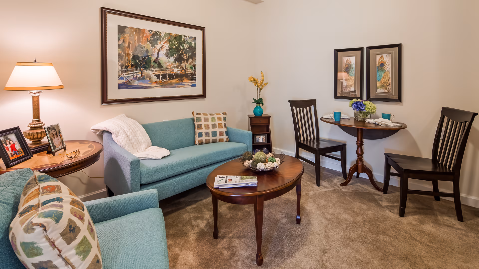 A cozy living room area with a teal sofa and armchair, a wooden coffee table with decorative items and a magazine, a side table with a lamp and framed photos, and a small round dining table with two chairs, set with cups and napkins. The walls have framed artwork and the floor is carpeted.