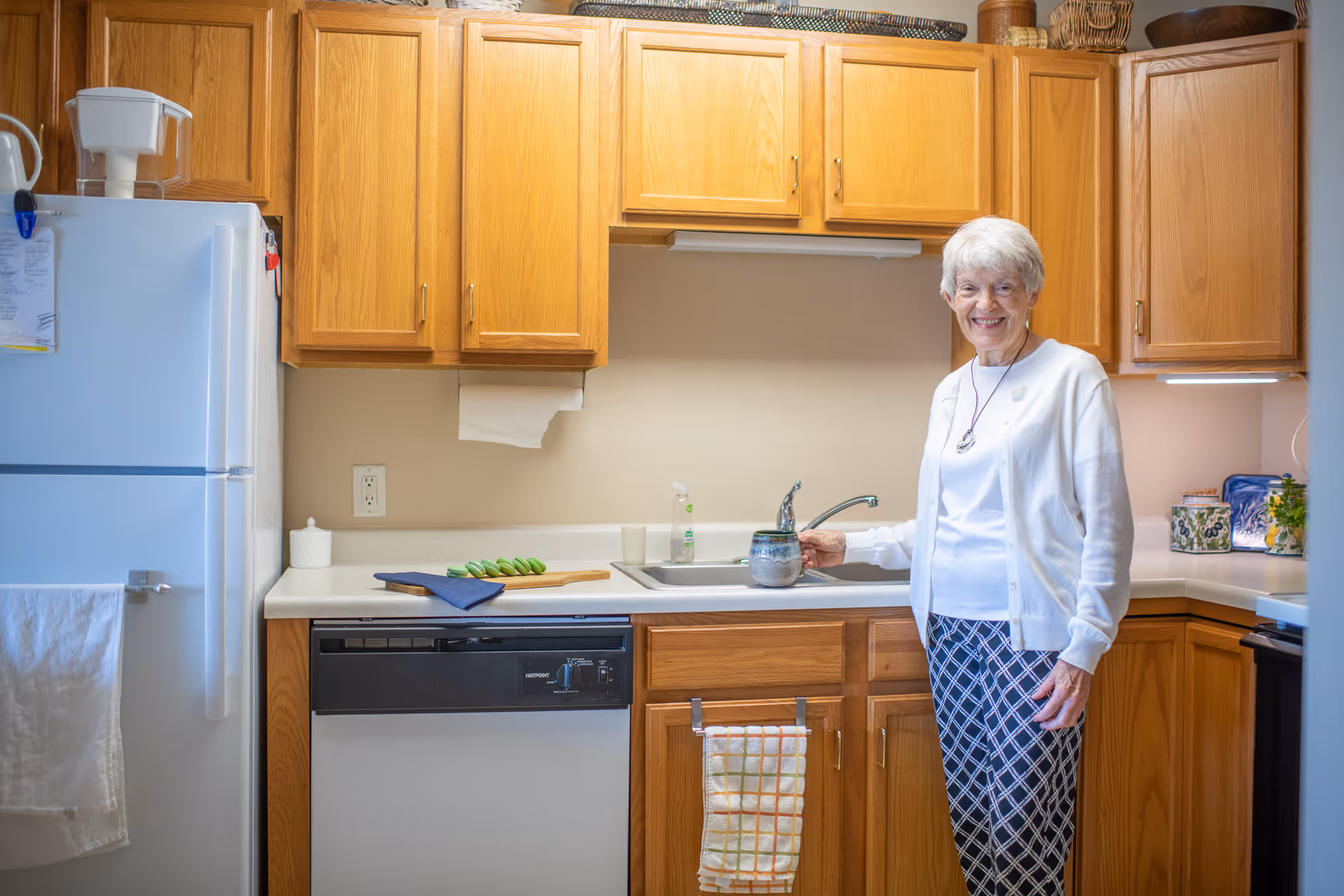 An elderly woman standing in a kitchen with wooden cabinets, a white refrigerator, a dishwasher, and a sink. She is smiling and holding a ceramic mug near the sink. The kitchen counter has a cutting board with green vegetables and a blue cloth.