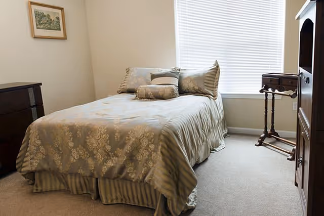 Small furnished bedroom with a neatly made bed, dresser, side table, and a window with blinds.