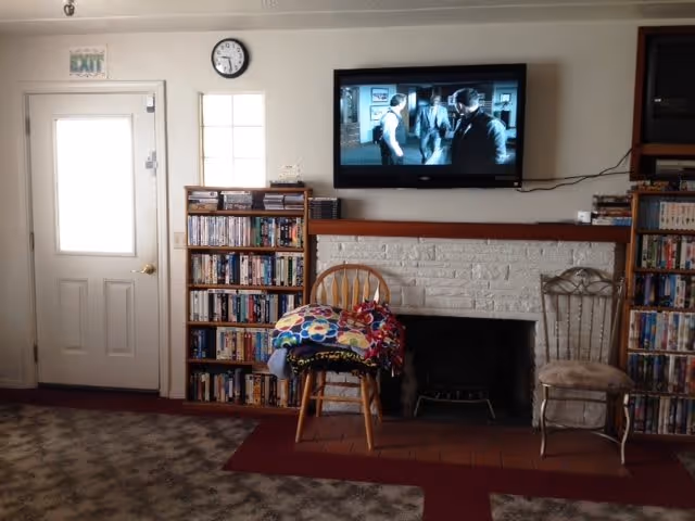 A cozy living room area with a white door on the left, a clock above a small window, and a flat-screen TV mounted on the wall showing a black and white scene. Below the TV is a white brick fireplace with a wooden mantel. Two chairs are placed in front of the fireplace, one wooden with colorful blankets draped over it and the other metallic with a cushioned seat. Bookshelves filled with DVDs or books flank the fireplace on both sides. The floor has a patterned carpet with a red rug near the fireplace.