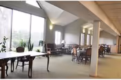A spacious, well-lit common area with large windows allowing natural light to fill the room. The room contains several tables and chairs arranged for seating, with some plants near the windows and wall-mounted lights providing additional illumination.