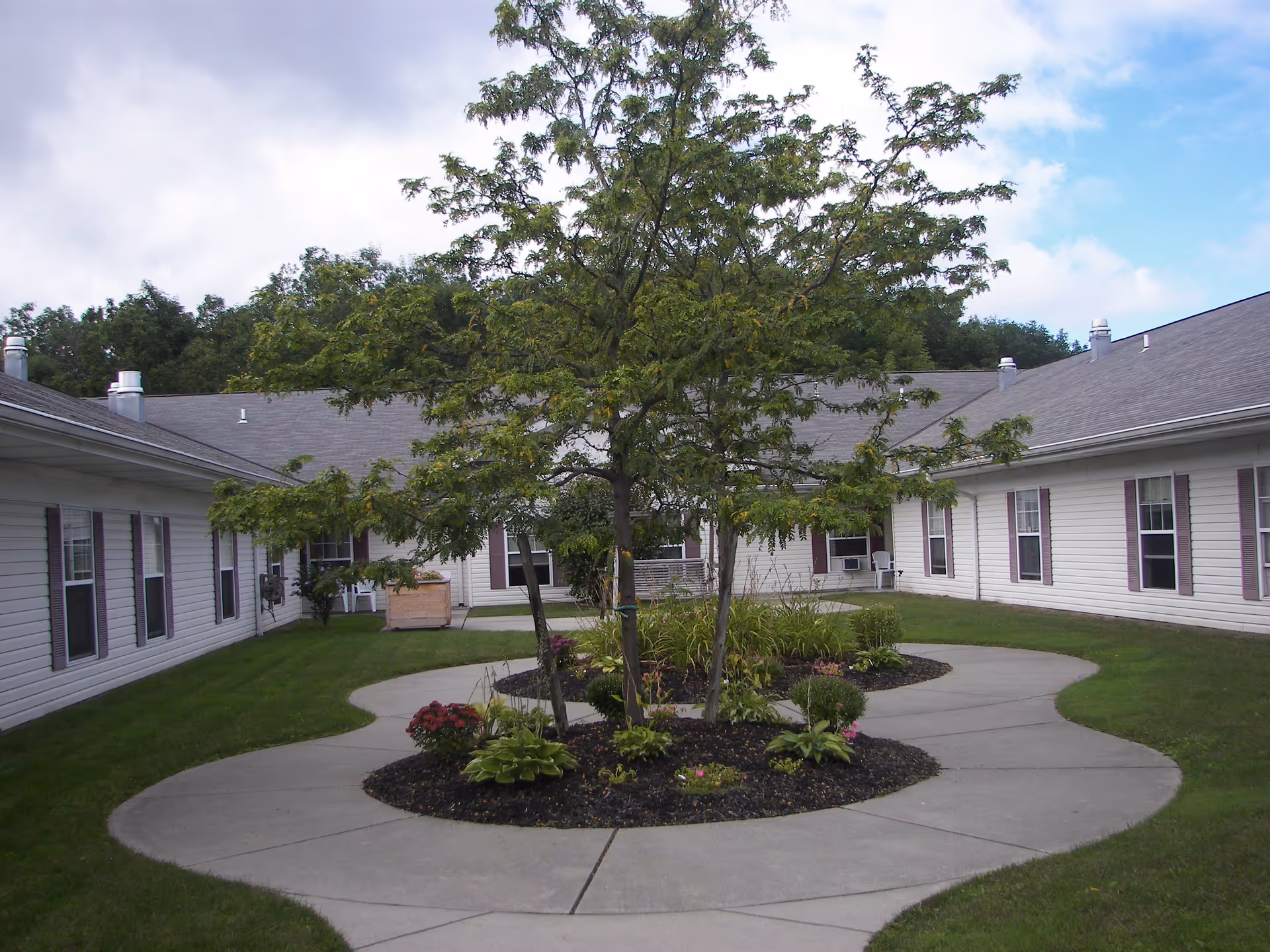 Outdoor courtyard area at Glenwell - A DePaul Senior Living Community featuring a circular concrete walkway surrounding a landscaped garden with trees and plants, bordered by a single-story building with white siding and multiple windows.