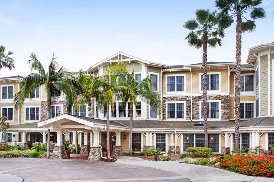 Front entrance of a multi-story senior living building with a covered porte-cochère, palm trees, and landscaped beds.