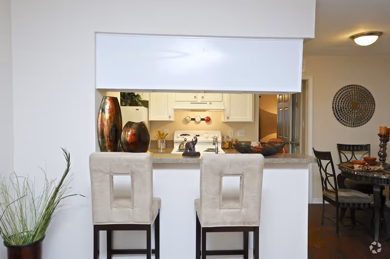 View of a kitchen pass-through with two bar stools at a counter, decorative vases, and a dining table visible to the right.