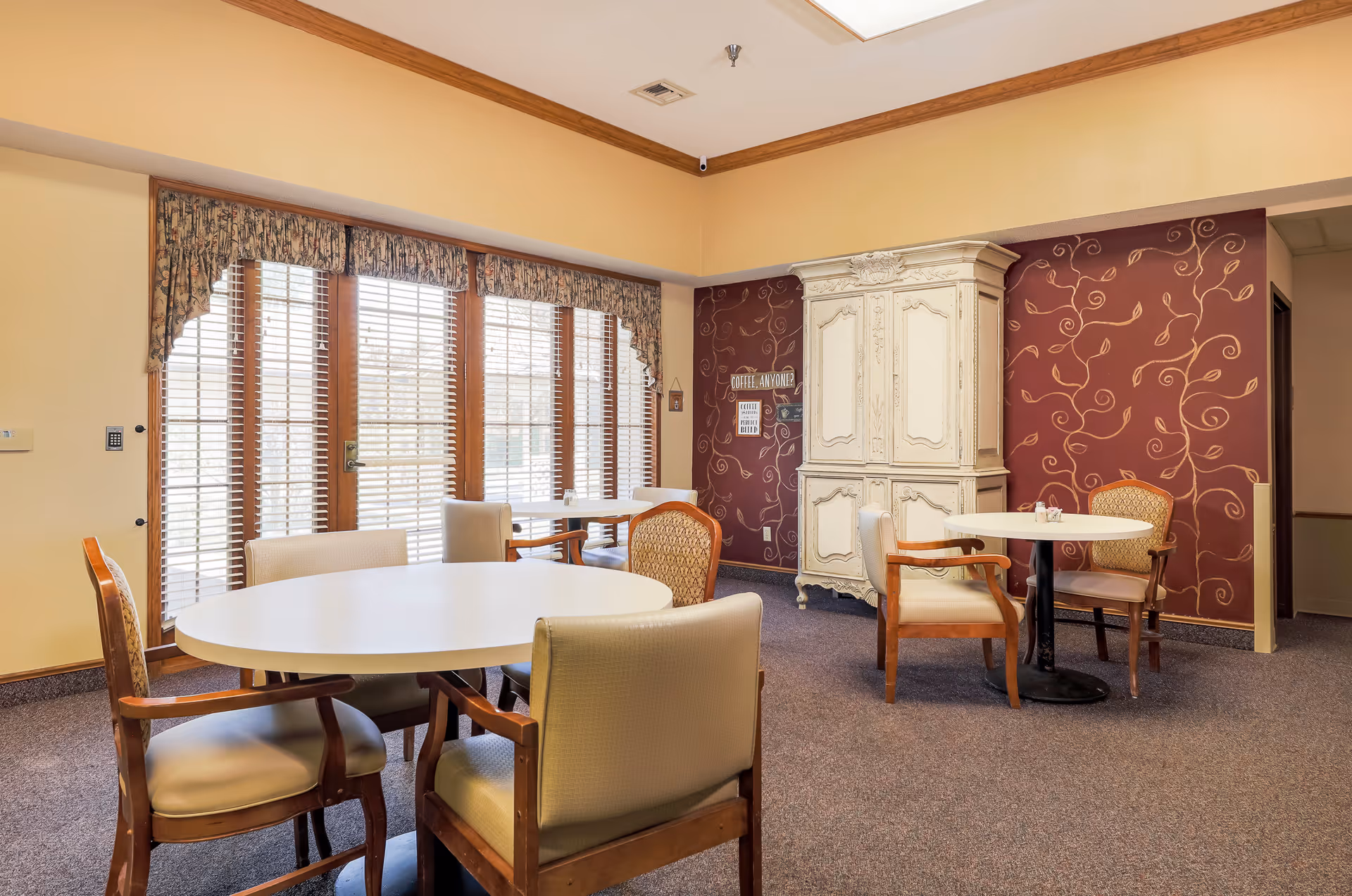 A cozy common area with round tables and cushioned chairs, large windows with wooden blinds and floral valances, a decorative white armoire against a maroon wall with gold vine patterns, and soft beige walls with wooden trim.