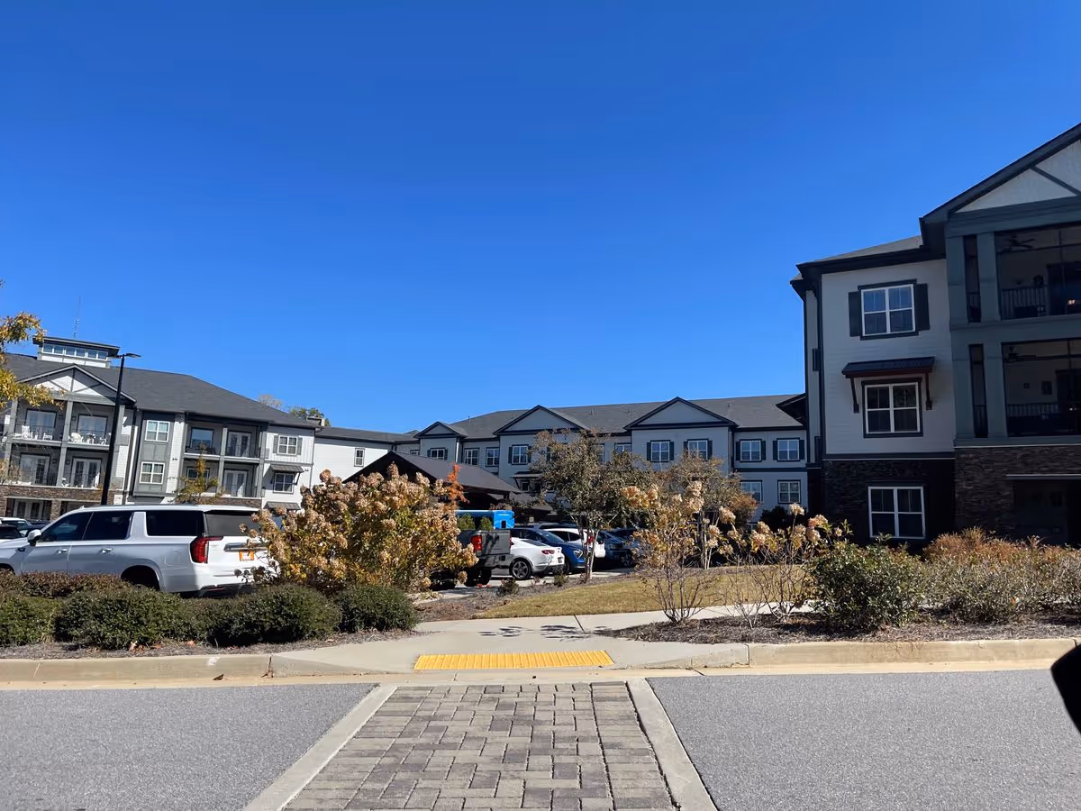 Exterior view of a multi-story senior living building with parked cars, landscaping, and a clear blue sky.