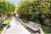 A paved walkway in a garden area lined with green trees and bushes on both sides, with a wooden bench on the right side of the path under the shade of the trees.