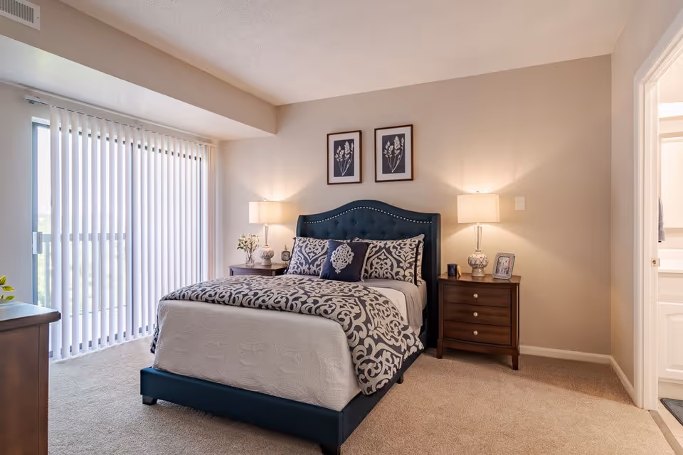 A neatly arranged bedroom with a blue upholstered bed featuring patterned bedding and pillows. There are two wooden nightstands on either side of the bed, each with a lamp and decorative items. Two framed botanical prints hang above the bed. Vertical blinds cover a large window or sliding door on the left side, allowing natural light to enter the room. The walls are painted beige and the floor is carpeted.