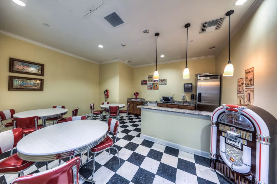 A retro-style dining area with black and white checkered flooring, several white oval tables surrounded by red and white chairs, a counter with a granite top, pendant lights hanging from the ceiling, a jukebox, and vintage-style wall decorations.