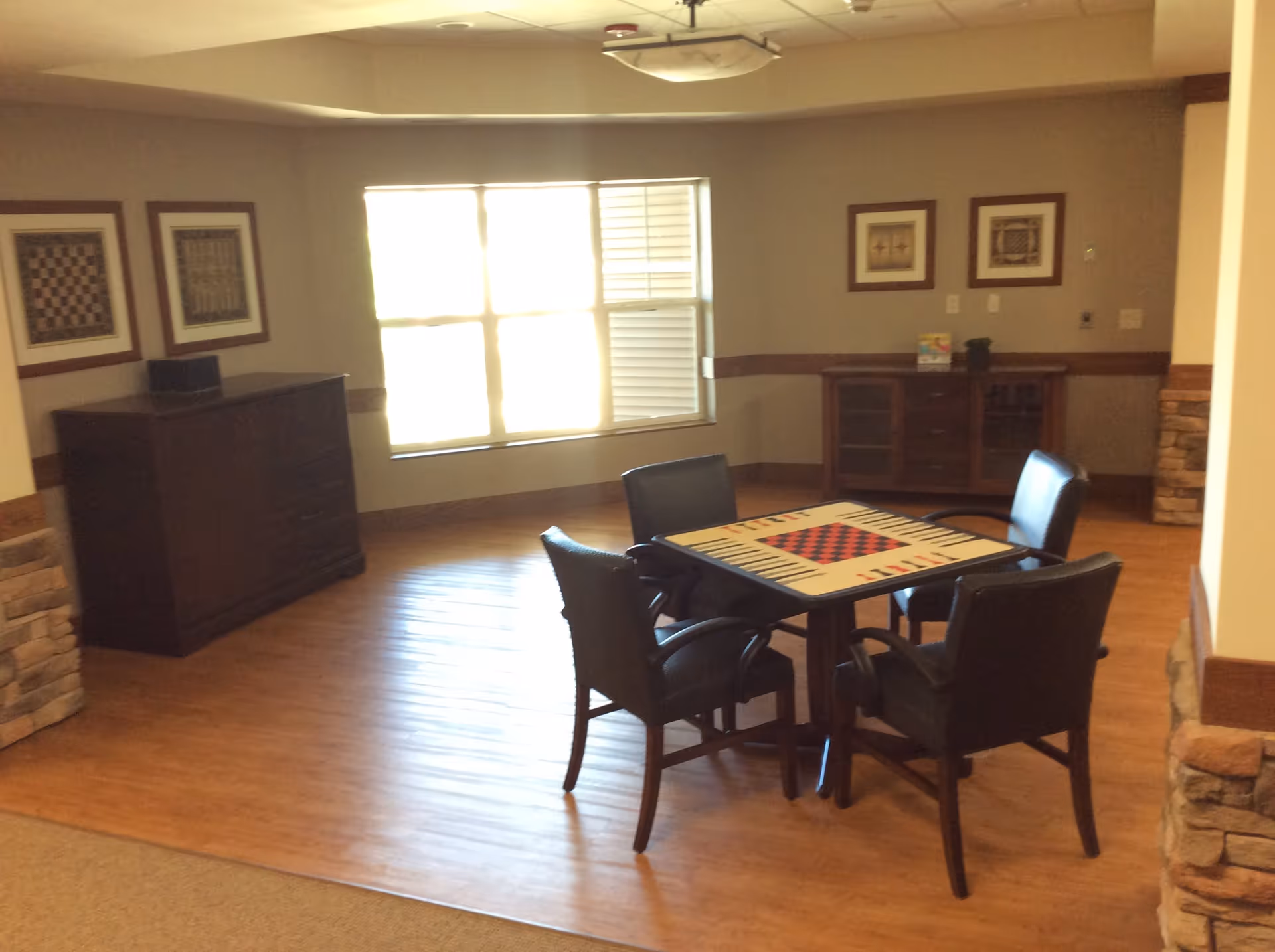 A well-lit room with wooden flooring featuring a square table with a checkerboard pattern on top, surrounded by four black chairs. The room has a large window letting in natural light, two wooden cabinets against the walls, and framed artwork hanging above the cabinets. The walls are painted beige with a wooden trim and stone accents on the corners.
