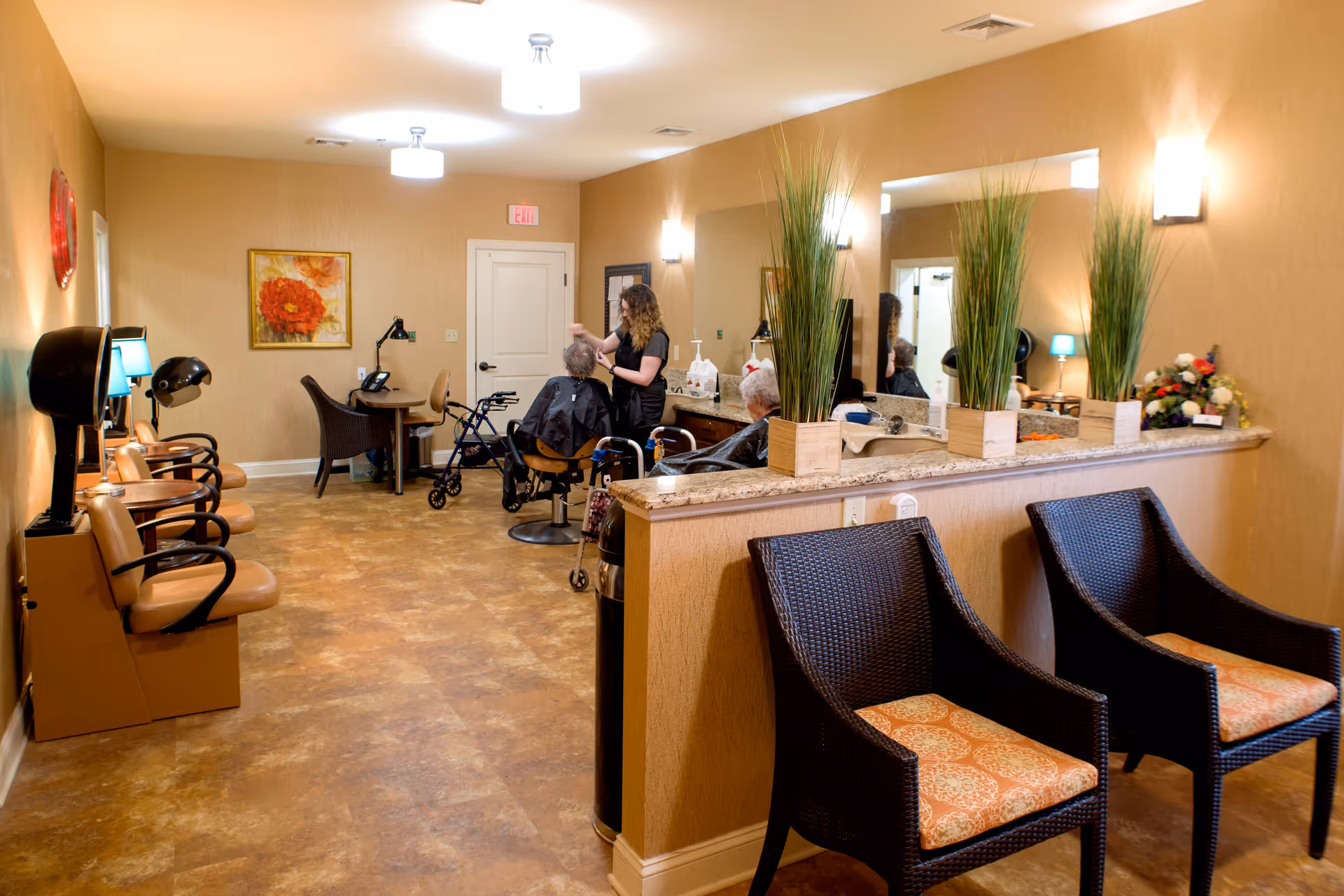 Interior of a salon area in a senior living facility with a stylist attending to an elderly woman seated in a salon chair. The room has warm beige walls, a large mirror, plants on a counter, wicker chairs with patterned cushions, and salon equipment including hair dryers and lamps.