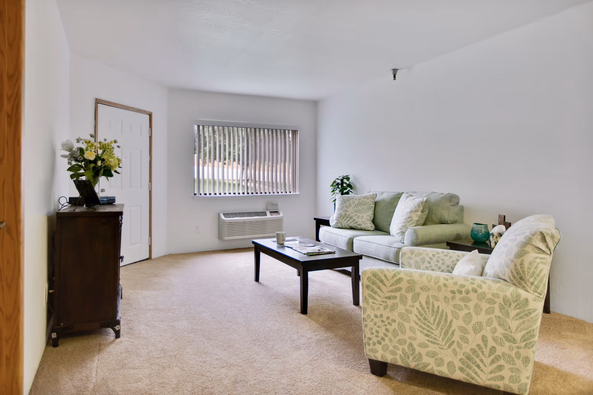 A bright living room with beige carpet, a light green sofa with patterned cushions, a matching armchair with leaf designs, a dark wooden coffee table with a cup and magazines, a small side table with a green vase and tissue box, a window with vertical blinds, and a wooden cabinet with a flower arrangement near a white door.