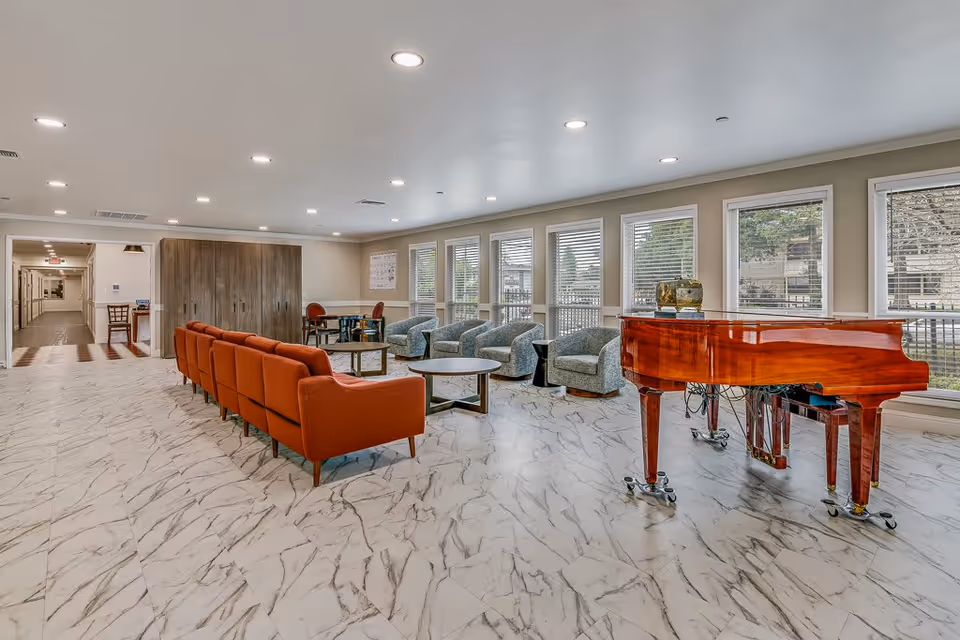 A spacious, well-lit common area in an assisted living facility featuring a row of orange armchairs, several gray swivel chairs, round tables, and a polished wooden grand piano near large windows with white blinds. The floor has a white and gray marble pattern, and there is a hallway visible in the background.