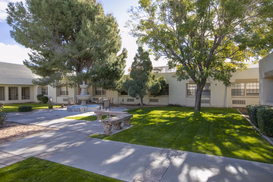 Outdoor courtyard area with green grass, trees, a stone bench, a multi-tiered water fountain, and several chairs and tables. The courtyard is surrounded by a single-story building with multiple windows.