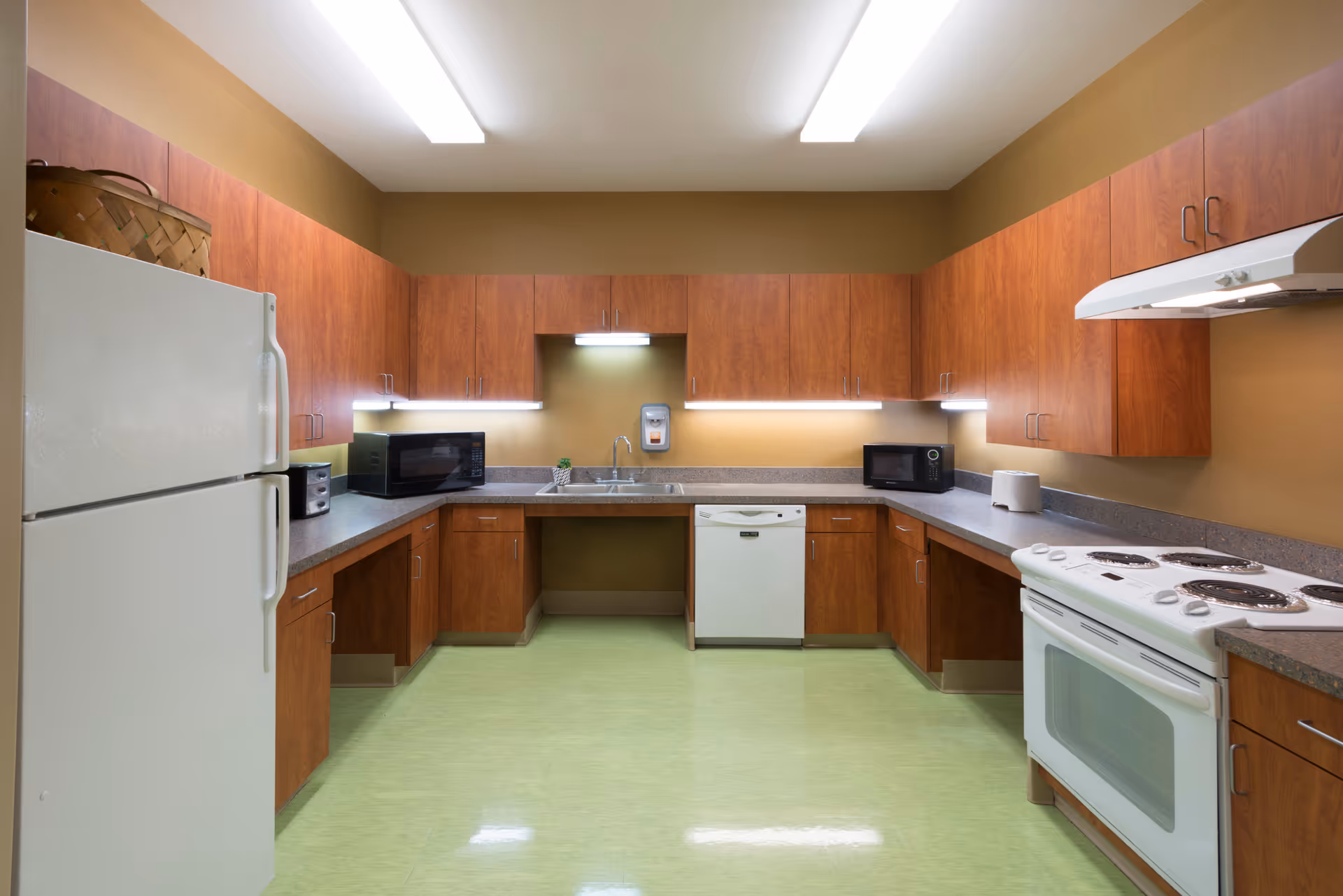 A clean and well-lit kitchen with wooden cabinets, a white refrigerator, a white electric stove with oven, two microwaves, a dishwasher, a toaster, and a sink with a soap dispenser mounted on the wall above it. The floor is light green and the walls are painted brown.