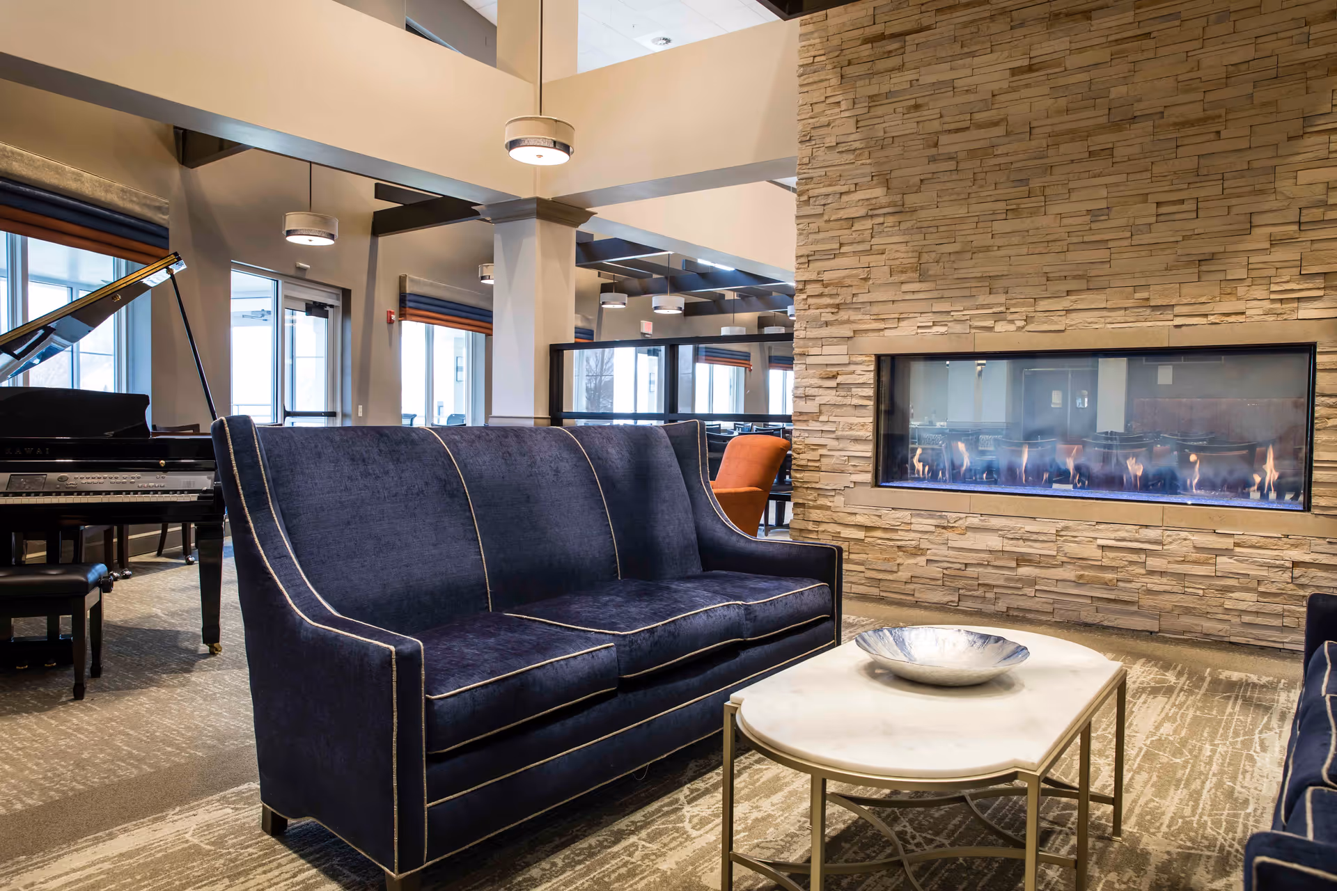 A modern living room area with a dark blue velvet sofa facing a rectangular gas fireplace set in a stone wall. A white marble coffee table with a decorative silver bowl is in front of the sofa. To the left, there is a black grand piano with a matching bench. The room has large windows and high ceilings with hanging round light fixtures.