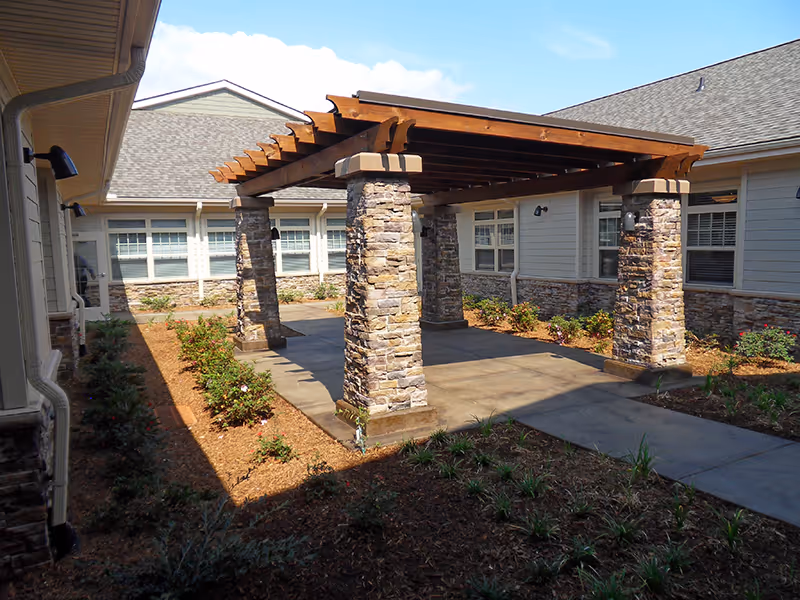Outdoor courtyard area with a wooden pergola supported by stone pillars, surrounded by a garden bed with small plants and shrubs. The courtyard is enclosed by a single-story building with multiple windows and a shingled roof under a clear blue sky.