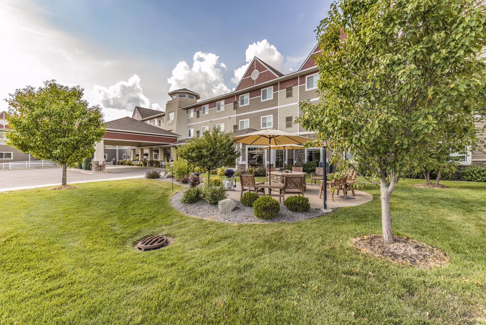 Front exterior of a senior living facility with a covered entrance, outdoor seating area with umbrella, and landscaped lawn and trees.