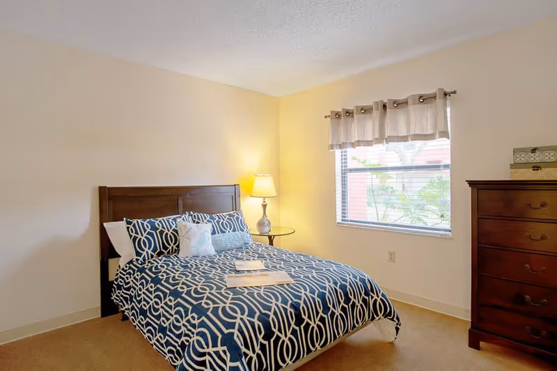 A bedroom with a bed featuring a blue and white patterned comforter and pillows, a wooden headboard, a round glass side table with a lamp, a window with beige curtains, and a wooden chest of drawers with decorative boxes on top.