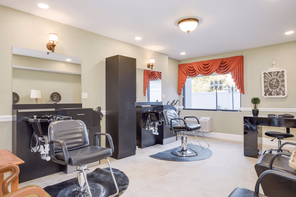 Interior view of a salon area in a senior living facility with two black salon chairs in front of wash basins and mirrors. The room has light-colored walls, a large window with red drapes, a wall clock, and a small black desk with a chair. The floor is tiled and the space is well-lit with ceiling lights.