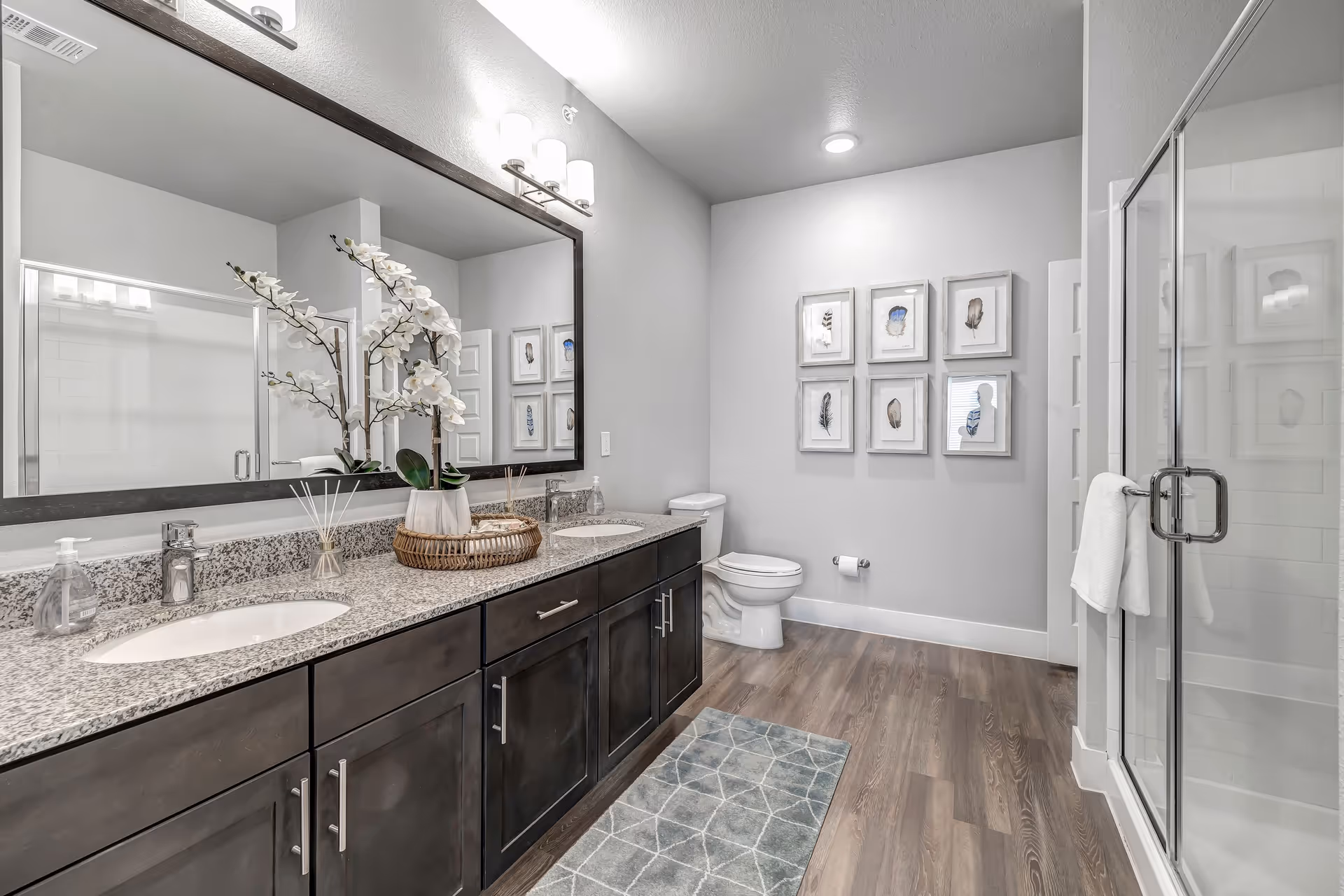 A modern bathroom with a double sink vanity featuring dark wood cabinets and a granite countertop. A large mirror with three light fixtures above it is mounted on the wall. There is a decorative tray with white orchids and reed diffusers on the countertop. The bathroom has a toilet, a glass-enclosed shower, framed artwork on the wall, and a patterned rug on the wooden floor.