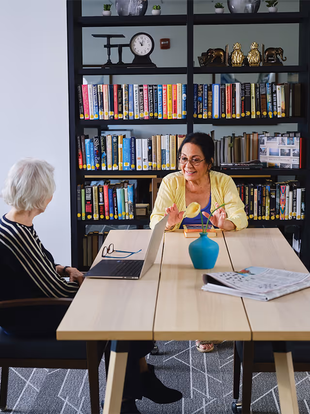 Two elderly women sitting at a wooden table in a library or reading room, engaged in conversation. One woman has white hair and is wearing a striped top, while the other has dark hair, glasses, and a yellow cardigan. A laptop, a pair of glasses, a blue vase with a yellow flower, and a newspaper are on the table. Behind them is a black bookshelf filled with books and decorative items including a clock and small sculptures.