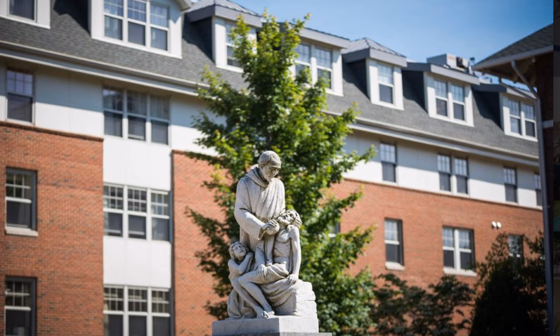 Stone statue of a robed figure with two children standing in front of a multi-story brick building and a leafy tree.