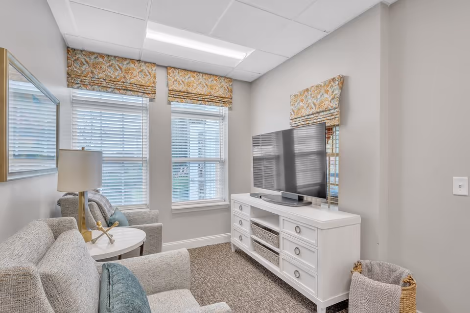 A cozy living room area with two gray upholstered armchairs, a small round white side table with a gold lamp, a white TV stand with drawers and baskets, and a flat-screen TV. The room has three windows with white blinds and patterned valances, beige walls, and carpeted flooring.