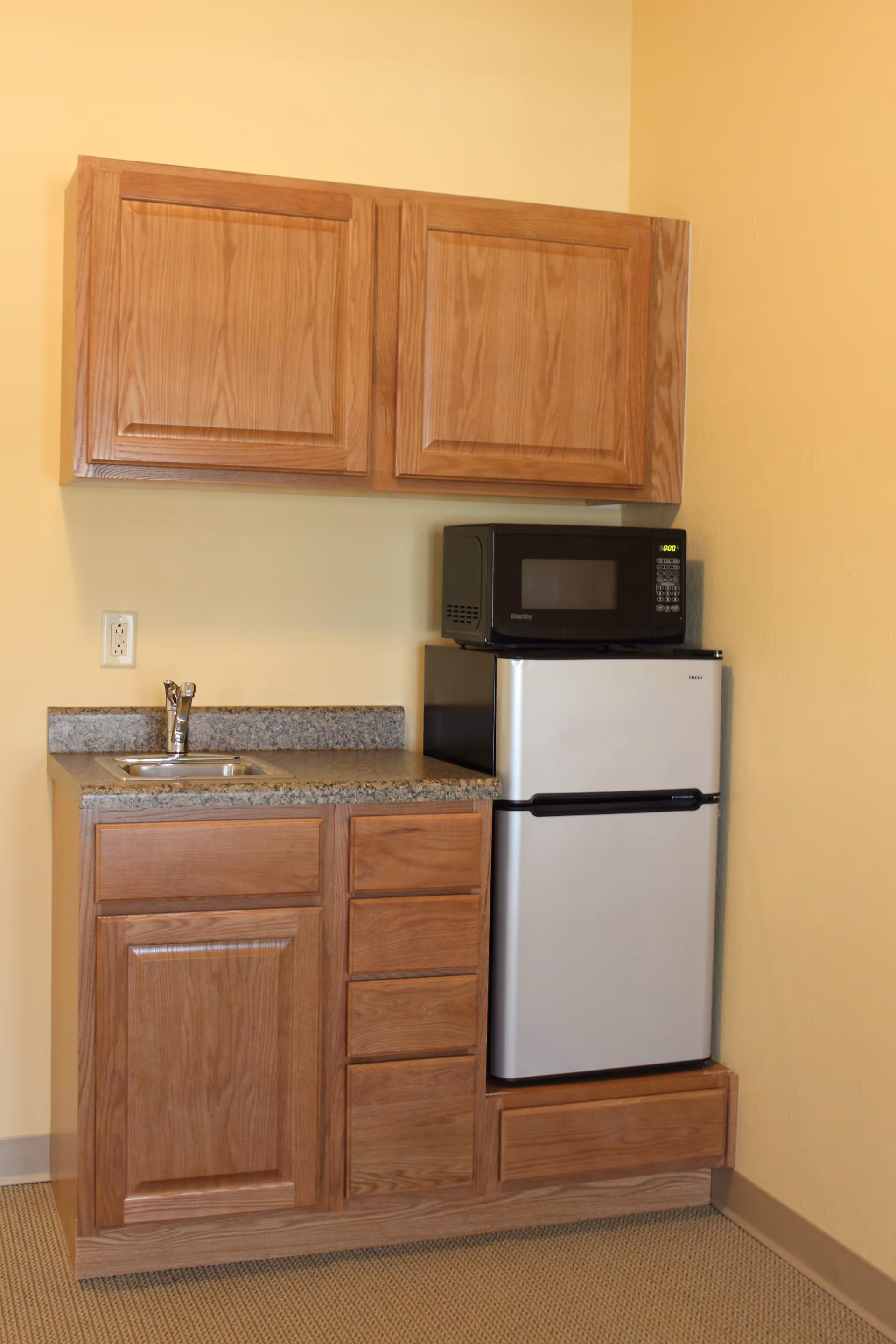 A small kitchenette with wooden cabinets, a granite countertop with a sink, a compact refrigerator, and a microwave placed on top of the refrigerator. The walls are painted light yellow and the floor is carpeted.