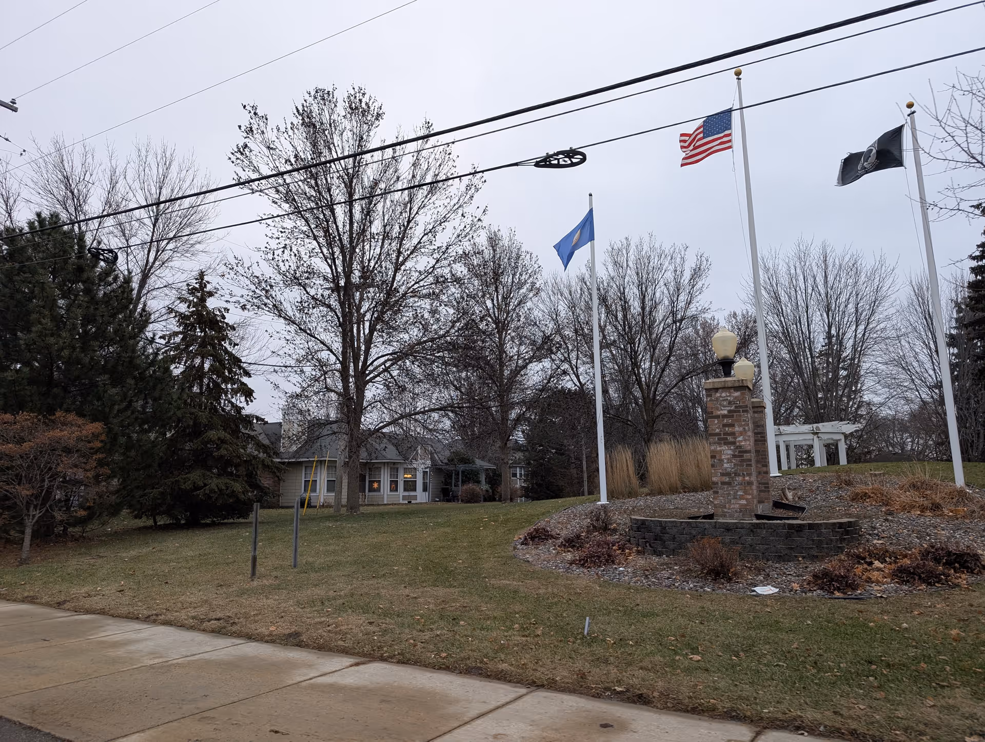 Exterior view of a building with flagpoles, a low brick monument, and leafless trees on a grassy lawn.