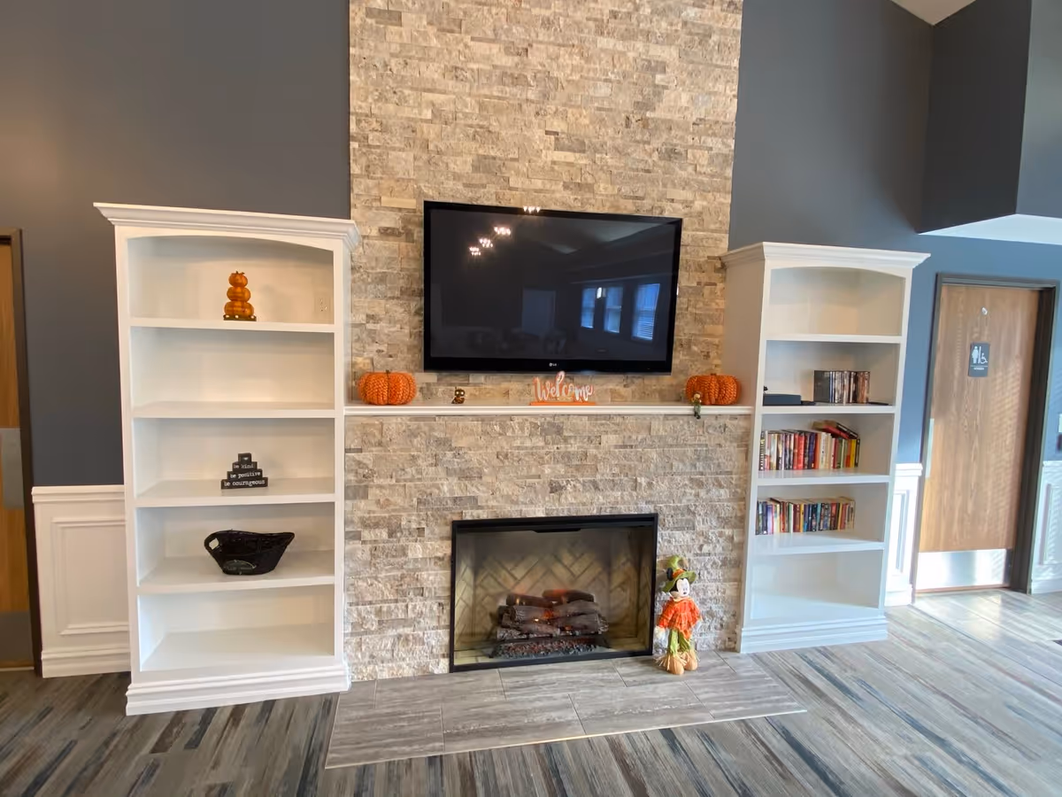 A cozy common area with a stone fireplace in the center, a flat-screen TV mounted above it, and two white bookshelves on either side. The shelves contain books, decorative pumpkins, and small ornaments. The floor has a wood-like finish, and there are two wooden doors visible in the background.