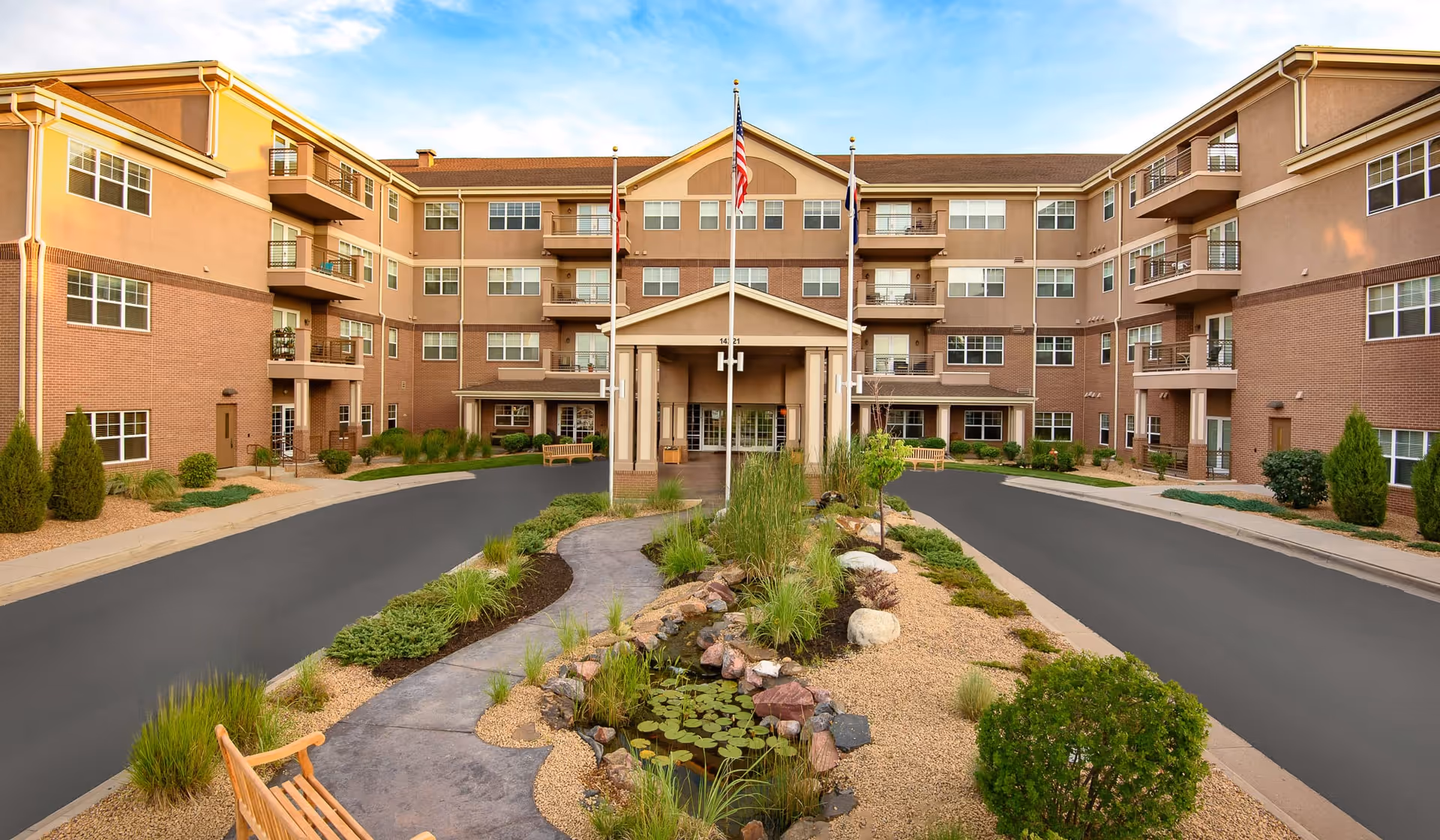Front exterior view of a multi-story senior living facility with a driveway leading to the entrance. The building has a combination of brick and beige siding with multiple windows and balconies. There are three flagpoles with flags in front of the entrance, surrounded by landscaped greenery, bushes, and a small water feature with rocks and plants. Wooden benches are placed along the pathway.