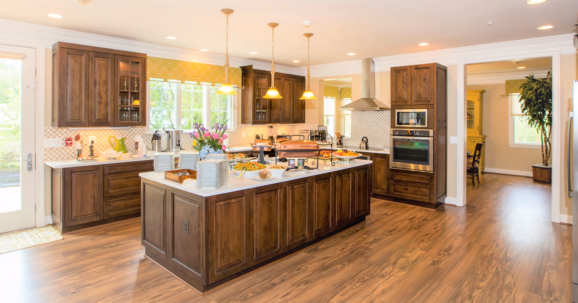 A spacious kitchen with wooden cabinets and a large central island topped with plates, flowers, and various dishes. The kitchen features stainless steel appliances, including a double oven, and has pendant lights hanging above the island. There is a window above the sink letting in natural light and a doorway leading to another room with a plant and a window.