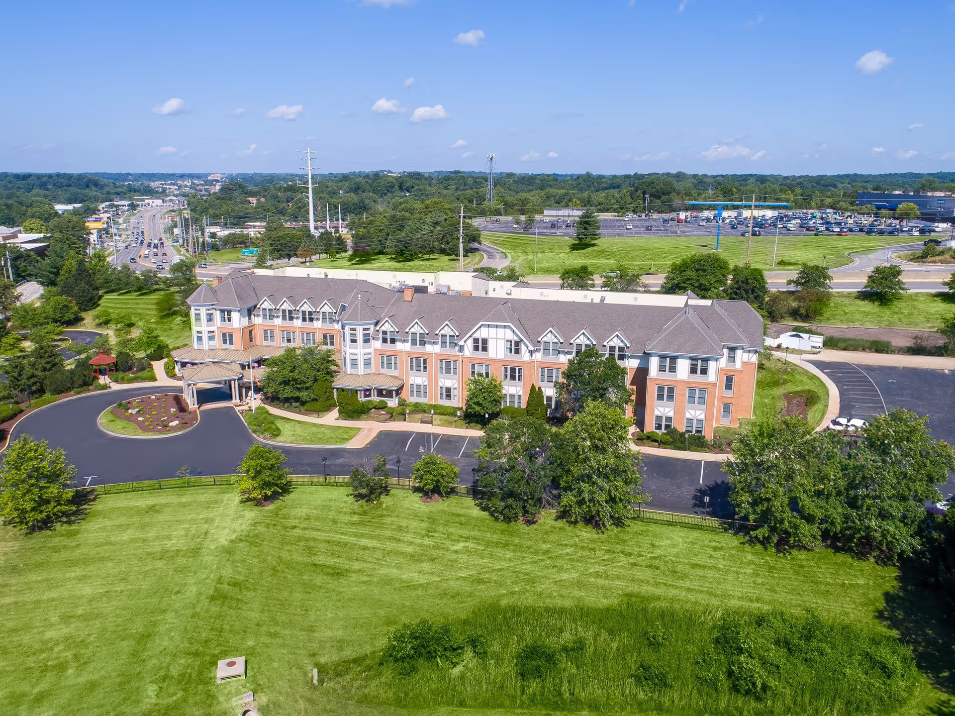 Aerial view of a large senior living facility building named Sunrise of Des Peres surrounded by green lawns, trees, and parking areas under a clear blue sky.