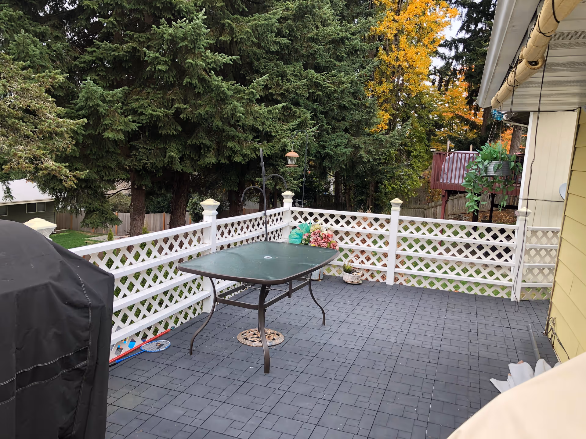 Outdoor patio area with a black metal table, white lattice railing, a covered grill, hanging plants, and trees in the background with some autumn foliage.