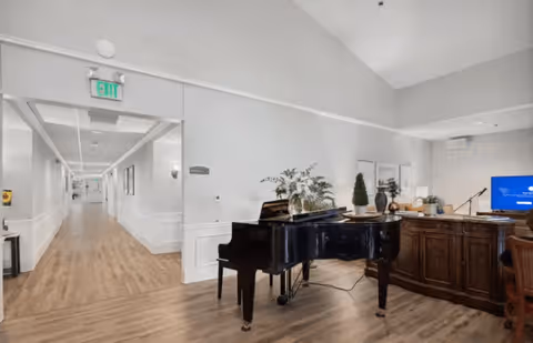 Interior view of a senior living facility hallway with light-colored walls and wood flooring. A black grand piano is positioned near a wooden reception desk adorned with decorative plants and a lamp. The hallway extends into the distance with framed artwork on the walls and an exit sign overhead.
