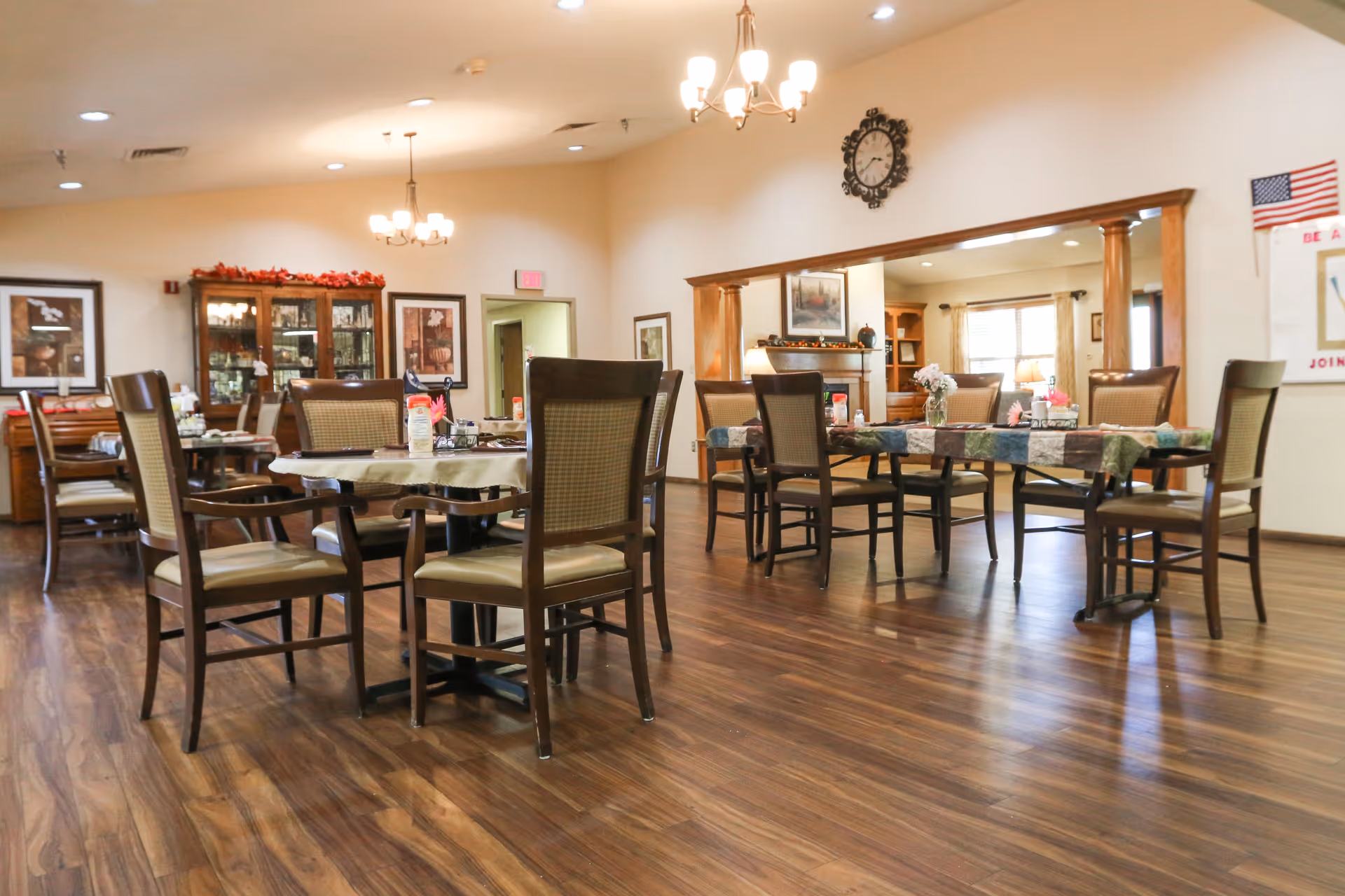 Spacious dining room with multiple round tables and chairs on wood floors, chandeliers, and a display cabinet.