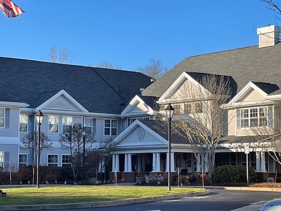 Exterior view of a large senior living facility building with white siding, multiple windows, and a covered entrance supported by white columns. There are leafless trees and lamp posts in front of the building, with a clear blue sky overhead.
