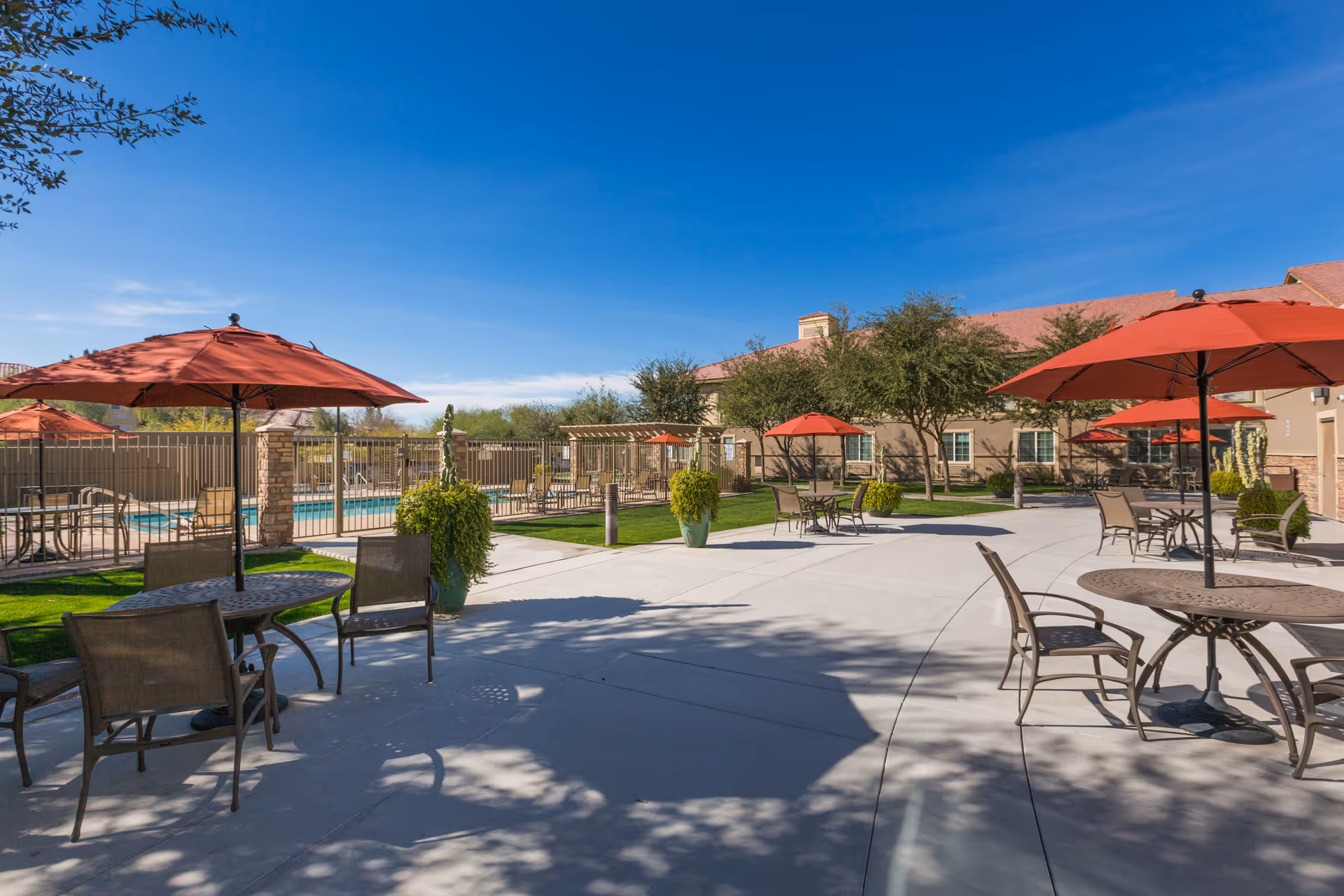 Outdoor patio area at Mountain Park Senior Living with several round tables and chairs under red umbrellas, a fenced swimming pool in the background, green grass, trees, and a clear blue sky.