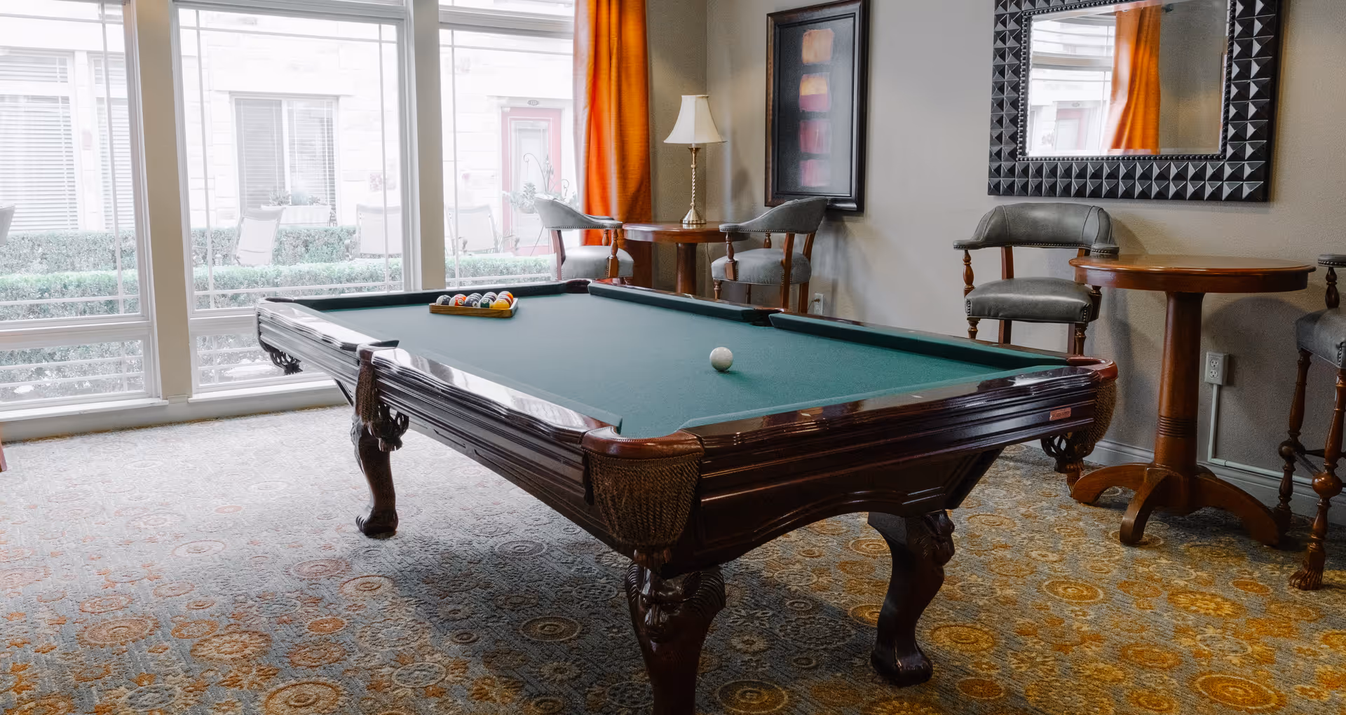 A room with a classic wooden pool table with green felt, a set of billiard balls arranged in a triangle, and a cue ball. The room has patterned carpet flooring, a large window with orange curtains, two wooden tables with chairs, a lamp, a framed artwork, and a decorative mirror on the wall.