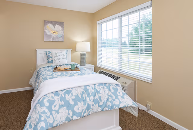 A cozy bedroom with a single bed featuring a white and blue floral comforter and a pillow with the text 'Good vibes only gorgeous'. A bedside table with a lamp is next to the bed. A large window with white blinds lets in natural light, and a floral painting hangs on the beige wall above the bed.