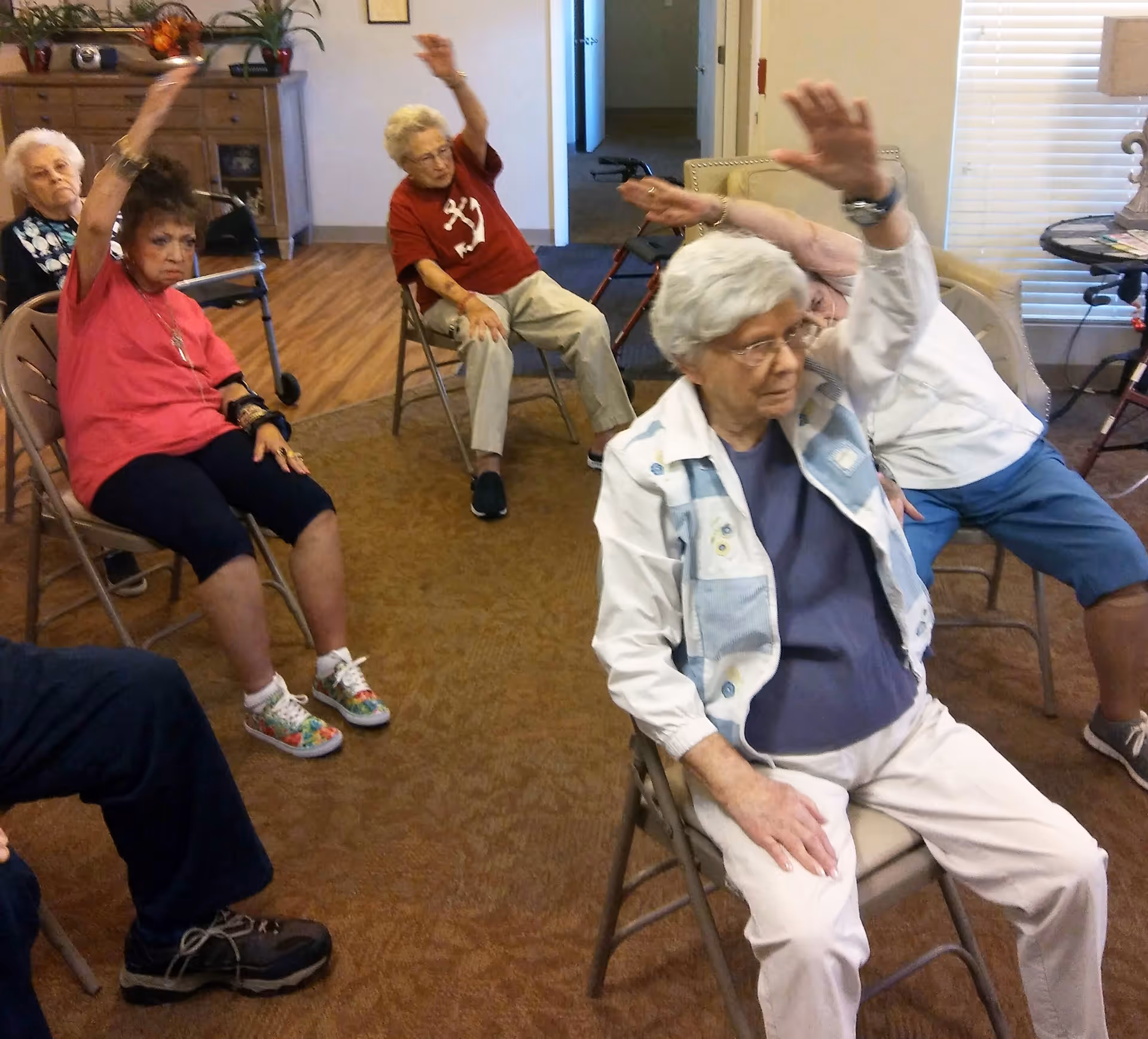A group of elderly individuals seated on chairs in a room, participating in a seated exercise session with their arms raised. The room has wooden flooring, a cabinet with plants and decorations, and a window with blinds.