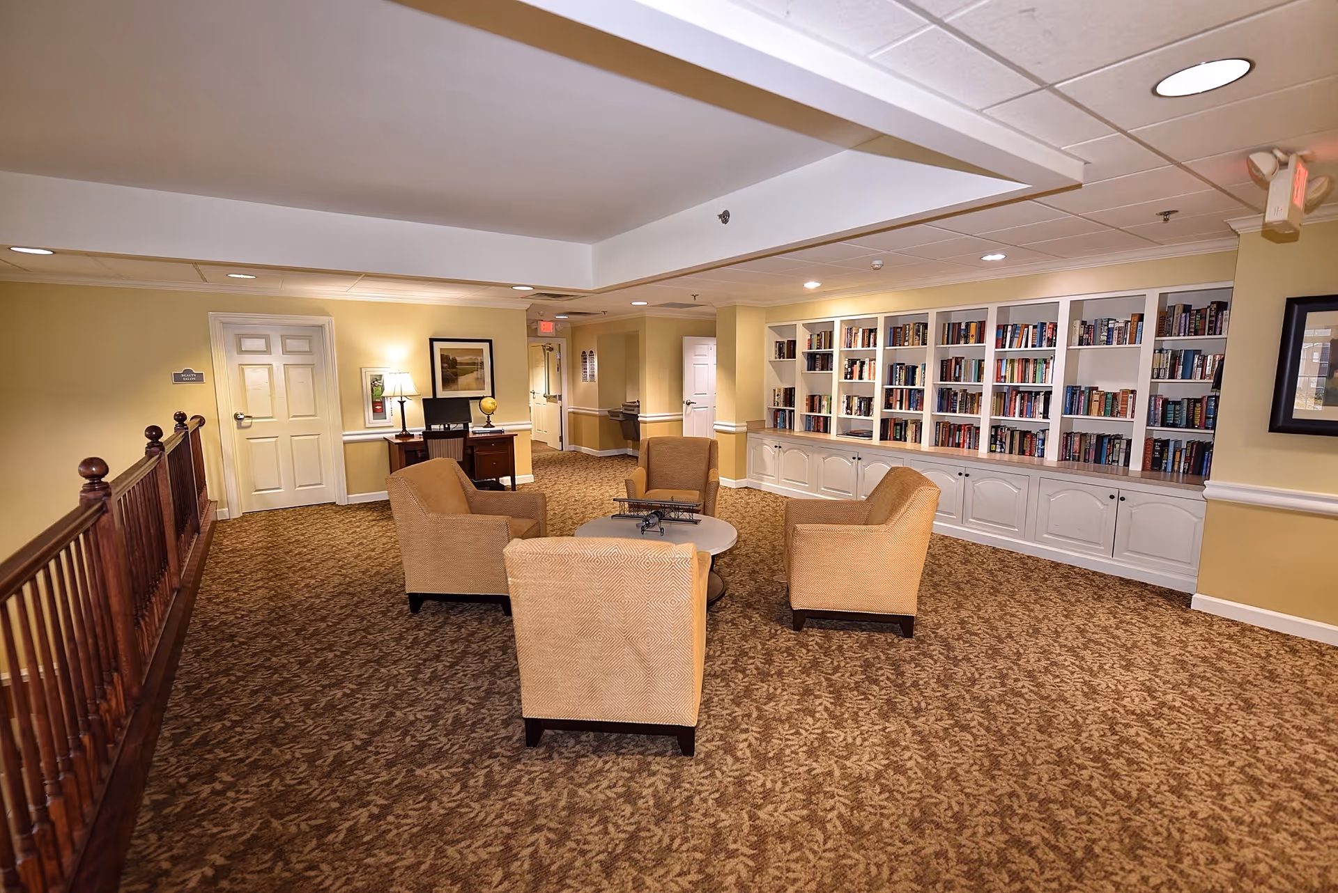 A cozy senior living facility common area with four beige armchairs arranged around a small round table. The room has a patterned carpet, yellow walls, and a large built-in white bookshelf filled with books. There is a wooden railing on the left side and a desk with a lamp and globe near a closed door in the background.